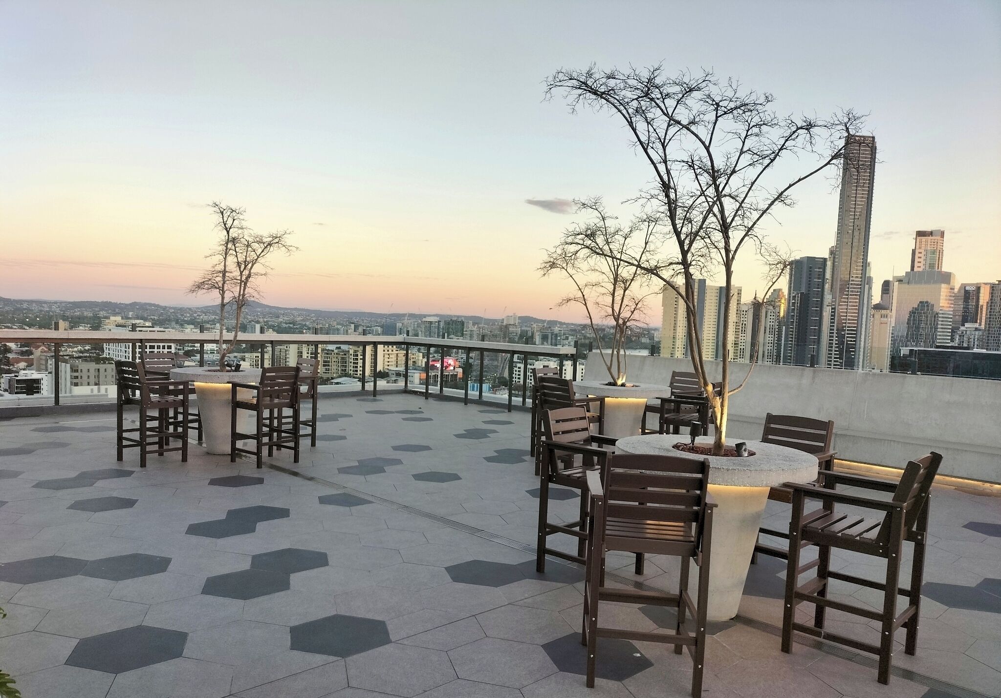 Eine Dachterrasse mit sechseckigen Fliesen, Holzstühlen und -tischen mit Blick auf eine Stadtlandschaft mit Gebäuden.