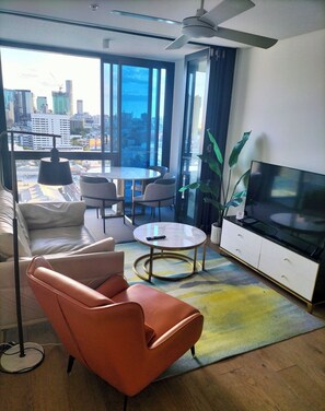 Ein modernes Wohnzimmer mit Sofa, Sessel, Esstisch, Fernseher und großen Fenstern mit Blick auf die Stadt.