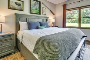 Una cama bien hecha con un edredón estampado, una mesita de noche con lámpara y una ventana con vistas a la vegetación.
