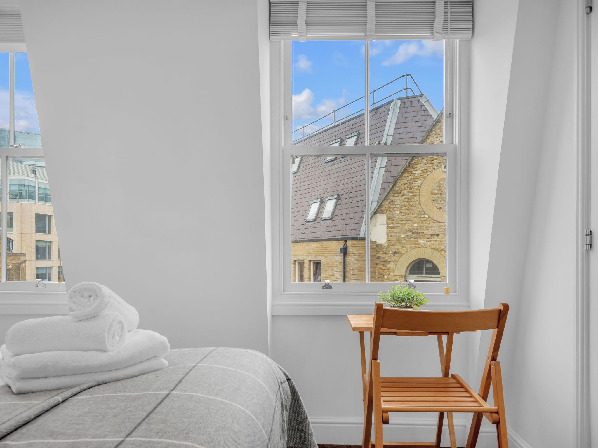 A bedroom with a bed, a chair, a window with a view of a building, and a stack of towels.
