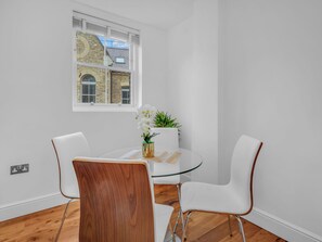 A modern dining area with a glass table, white chairs, and a view of a stone building through a window. - Spitafields Apartment by Interhome (Clerkenwell)