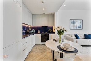 Eine moderne Küche mit weißen Schränken, ein Essbereich mit rundem Tisch und ein Wohnzimmer mit Sofa und einem gerahmten Bild an der Wand.