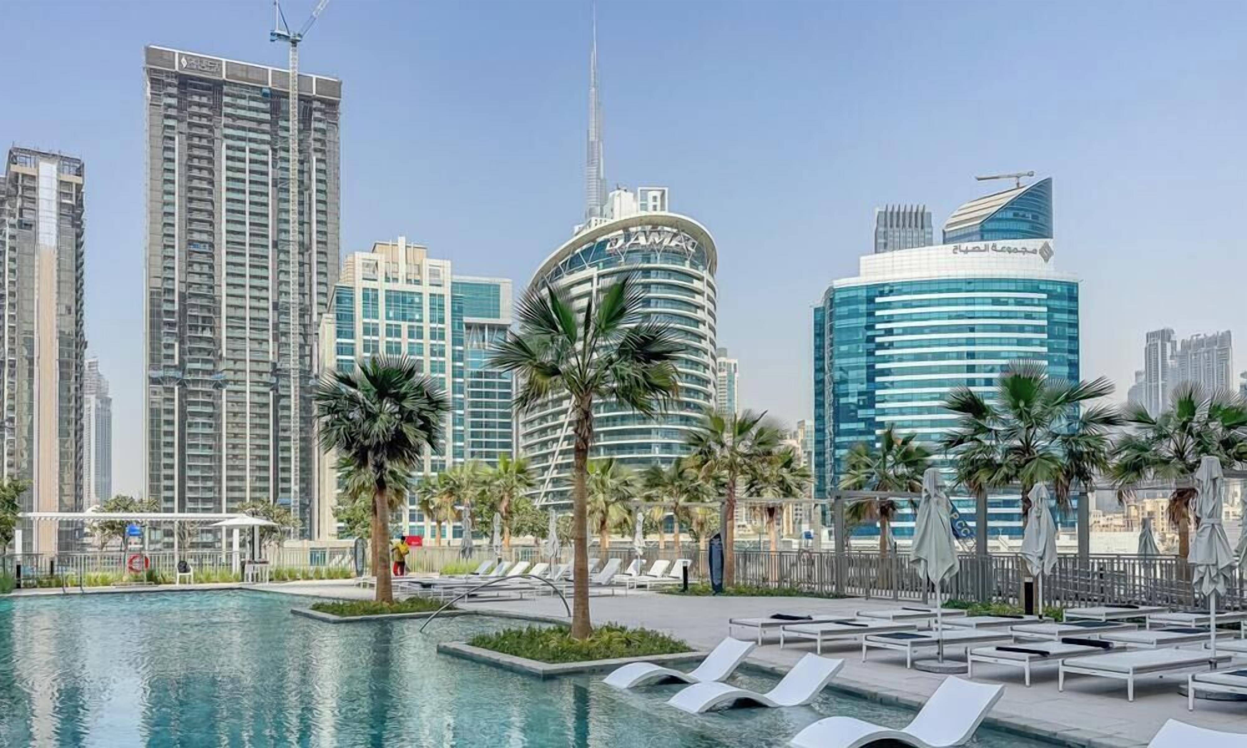 A poolside area with lounge chairs and palm trees, surrounded by high-rise buildings.