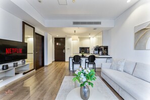 Un saló modern amb televisió plana, un sofà, una taula baixa i una zona de menjador amb cadires, i una cuina al fons.