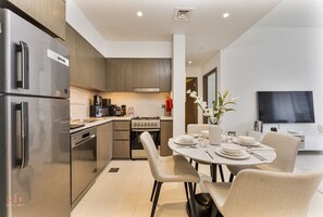 A modern kitchen with stainless steel appliances, a dining area with a round table and chairs, and a flat-screen TV mounted on the wall. - Madely | Chic Act One Haven | Burj Khalifa (Dubai)
