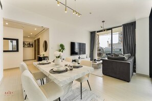 A modern living room with a sofa, a dining table set for four, and a flat-screen TV mounted on the wall. - Madely | Stylish 2BR with Private Pool (Dubai)