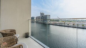 Un balcon avec des fauteuils en osier, une rambarde en verre et une vue sur une rivière, avec des bâtiments et des chantiers de construction en arrière-plan.