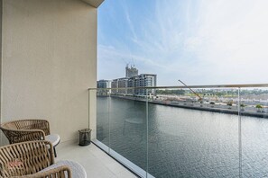 A balcony with wicker chairs, a glass railing, and a view of a river with buildings and construction in the background. - Canal-Side Living  Elegant 2BR in Canal Front (Dubai)