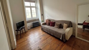 Ein Zimmer mit Holzboden, einem grauen Sofa mit Kissen, einem kleinen Tisch und einem Fenster mit Jalousien.