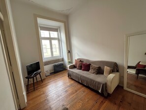 Ein Zimmer mit Holzboden, einem grauen Sofa mit Kissen, einem kleinen Tisch und einem Fenster mit Jalousien.