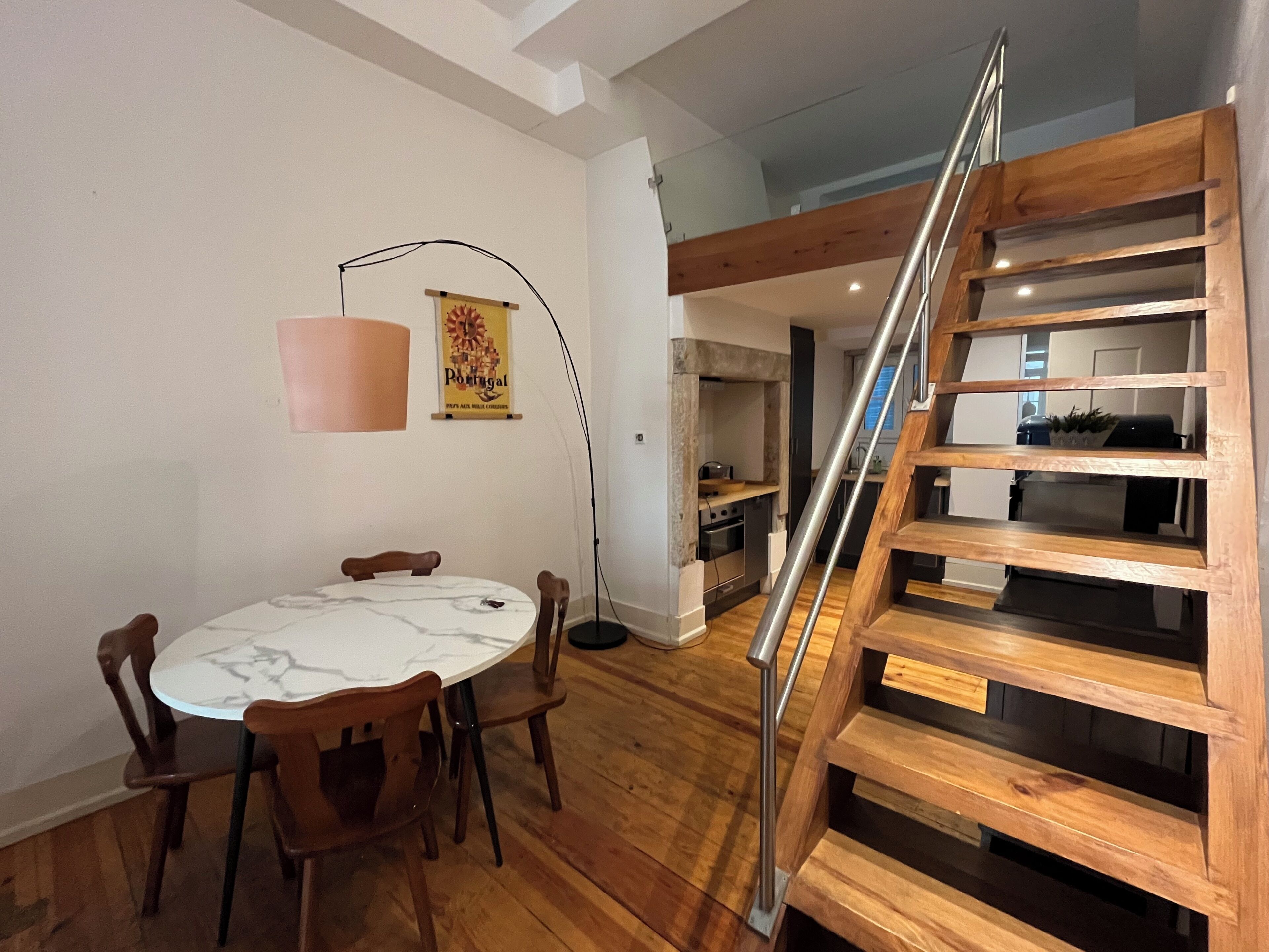 A dining area with a marble table and wooden chairs, a staircase with a metal railing, and a kitchen in the background.