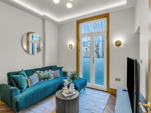 Ein modernes Wohnzimmer mit einem türkisfarbenen Sofa, einem runden Couchtisch, einem Flachbildfernseher und einem großen Fenster mit Blick auf Bäume.