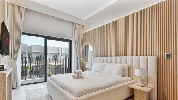 A modern hotel room with a large bed, a bedside table with a lamp, and a view of the city through a balcony. - Spacious | 4 Bed Villa | Private Pool | Meydan (Dubai)
