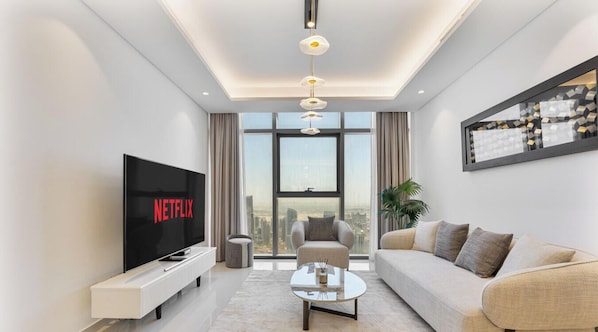 A modern living room with a large flat-screen TV, a sofa, a coffee table, and a city view through large windows. - High-Rise | 3 Bed Paramount Midtown | Burj View (Dubai)