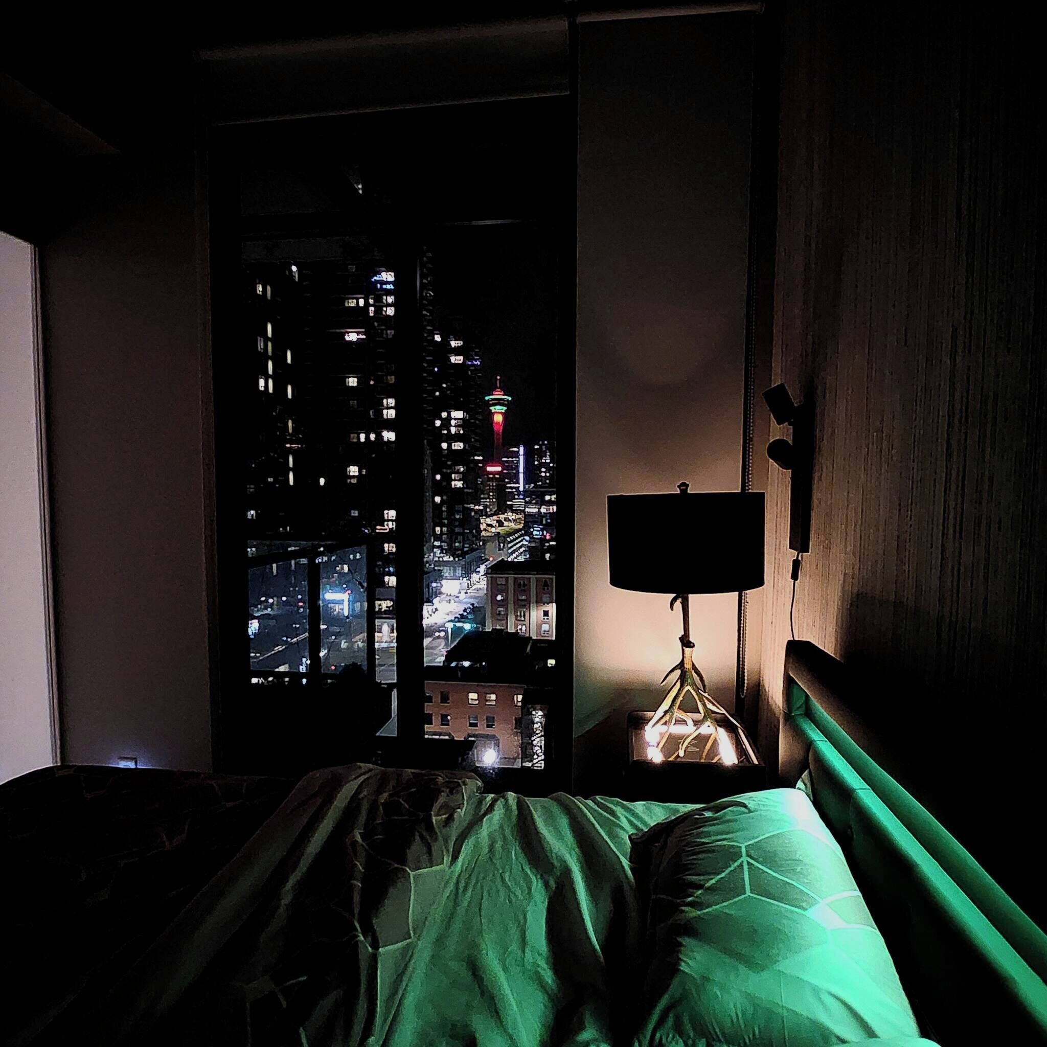 A bedroom with a view of a cityscape at night, a bedside table with a lamp, and a bed with green bedding.