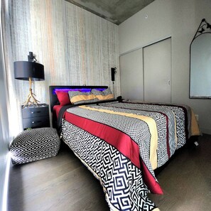 A bedroom with a patterned bedspread, a bedside lamp, and a wall-mounted light fixture. - Spectacular Tower View Condo w FREE parking n (Calgary)