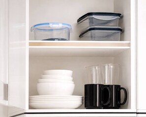 A kitchen cabinet with storage containers, white plates, and black mugs. - S&G Beachfront Serviced 1-BR, Step to Tops and 7-11 (Pattaya City)