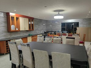 A modern dining area with a large table, chairs, wooden cabinets, and a kitchen in the background.