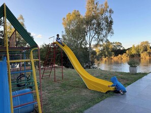 Un parc infantil amb un tobogan groc, una estructura per a escalar i una massa d'aigua al fons.