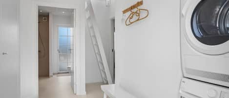 Une buanderie avec des machines à laver superposées, un banc et une œuvre d’art en bois accrochée au mur.