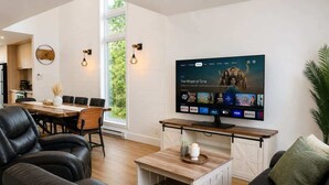 A modern living room with a flat-screen TV displaying a streaming service interface, a dining area with a wooden table and chairs, and a large window providing natural light. - Chic 2-story condo, a nature retreat for 6 (Québec City)