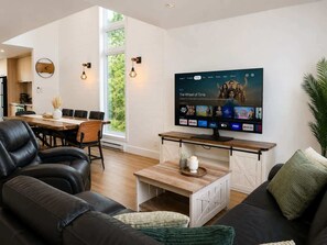 Un salon moderne doté d’un téléviseur à écran plat affichant l’interface d’un service de diffusion en continu, d’un coin repas avec une table et des chaises en bois, et d’une grande fenêtre laissant entrer la lumière naturelle.
