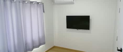 Et soveværelse med en seng, et fladskærms-tv på væggen, en klimaanlæg og et vindue med gardiner.