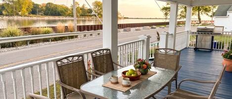 A covered outdoor patio with a table set for two, chairs, a grill, and a view of a road and water.