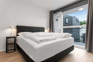 Ein modernes Schlafzimmer mit einem großen Bett, zwei Nachttischlampen und Blick auf ein Gebäude durch ein großes Fenster.