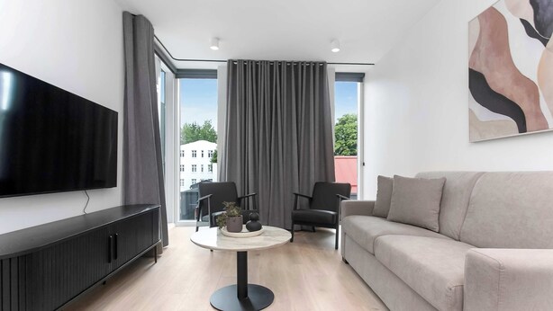 Een moderne woonkamer met een flatscreen televisie, een ronde salontafel, een bank en een groot raam met gordijnen.
