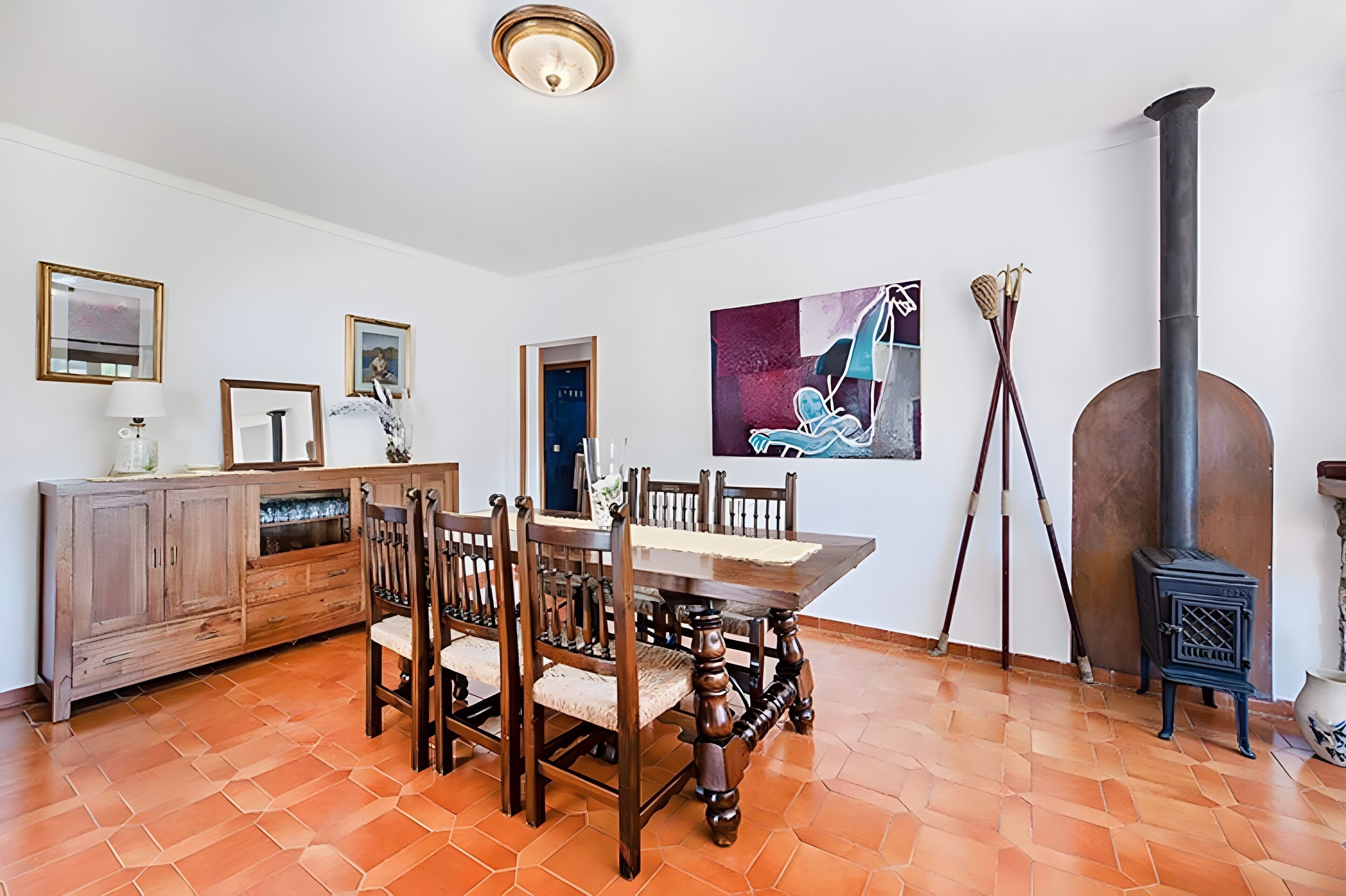 A dining area with a wooden table and chairs, a cabinet, a painting, and a wood-burning stove.