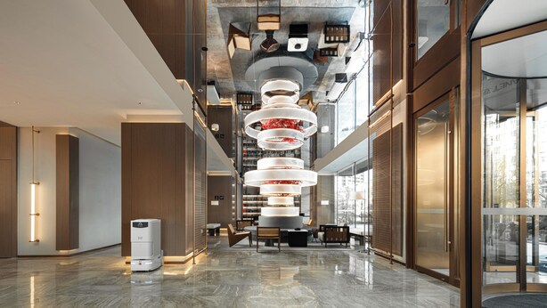 Een moderne lobby met een centraal, rondom lichtarmatuur, een receptiebalie en een glazen ingang.