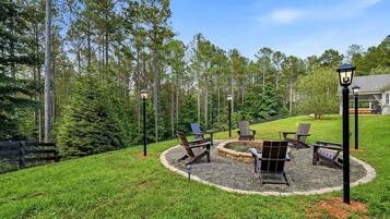 A circular outdoor seating area with chairs and a fire pit, surrounded by a gravel ring, set against a backdrop of tall trees and a grassy lawn.