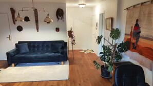 Un salon avec un canapé bleu foncé, un plancher en bois, une plante en pot, un fauteuil de bureau noir et une tapisserie colorée accrochée au mur.