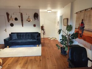 Ein Wohnzimmer mit dunkelblauem Sofa, Holzboden, einer Topfpflanze, einem schwarzen Bürostuhl und einem farbenfrohen Wandteppich.
