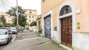 Una calle estrecha con coches estacionados, un edificio con una puerta marrón y un ventanal con reja, y otro edificio al fondo.