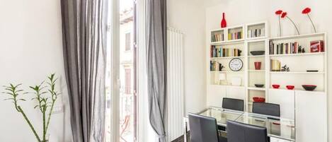 A modern dining area with a glass table, black chairs, a white bookshelf with books and decorative items, and a view through a sliding glass door.