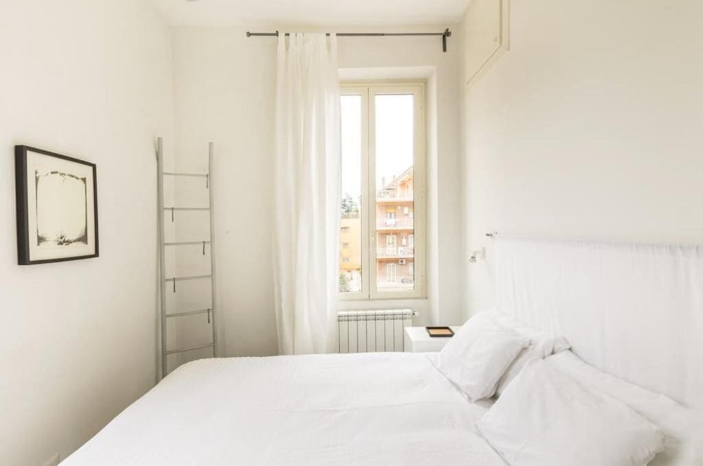 A bedroom with a bed, a window with curtains, a radiator, and a framed picture on the wall.