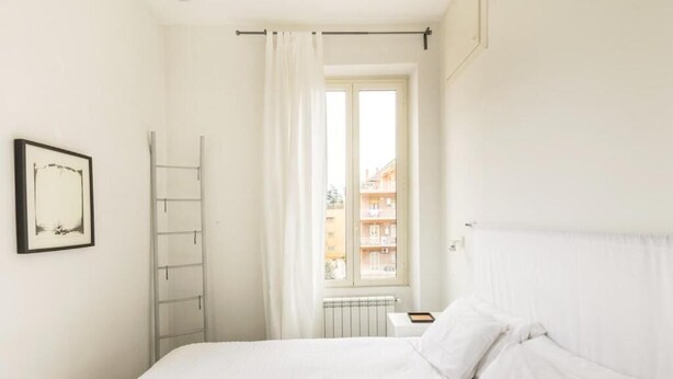 Un dormitorio con cama, ventana con cortinas, radiador y un cuadro en la pared.