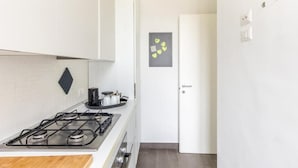 Una cocina con estufa a gas, un hervidor negro y una pequeña estantería con utensilios de cocina. Se observa una puerta blanca y una obra de arte gris en la pared de encima.