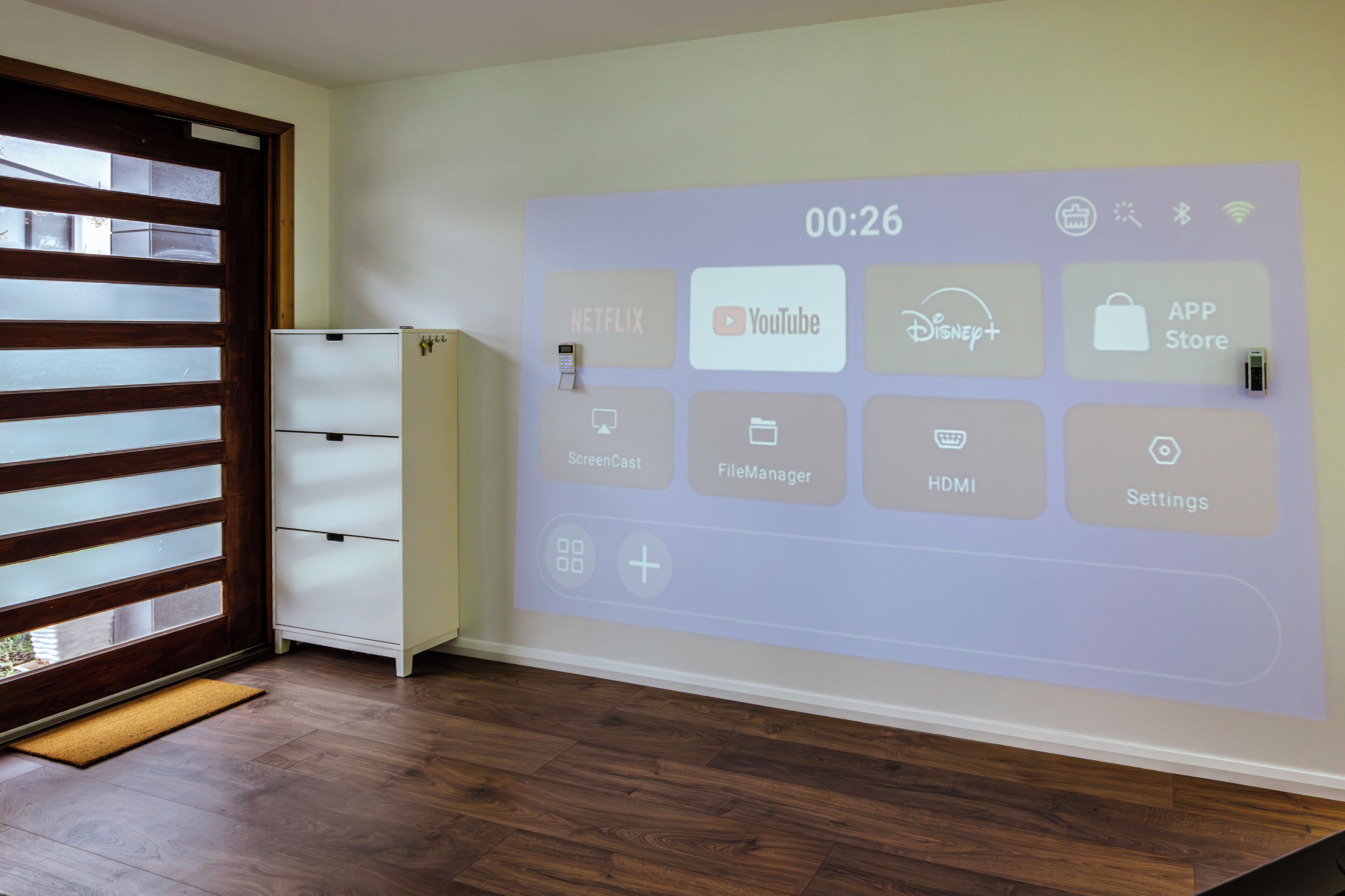 A room with a wall-mounted projection displaying various app icons, a white cabinet, and a wooden floor.