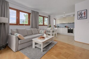 A modern living room with a grey sofa, a coffee table with pastries, and a dining area with white chairs. - Lumina 3-room Apart with Parking  (Warszawa)
