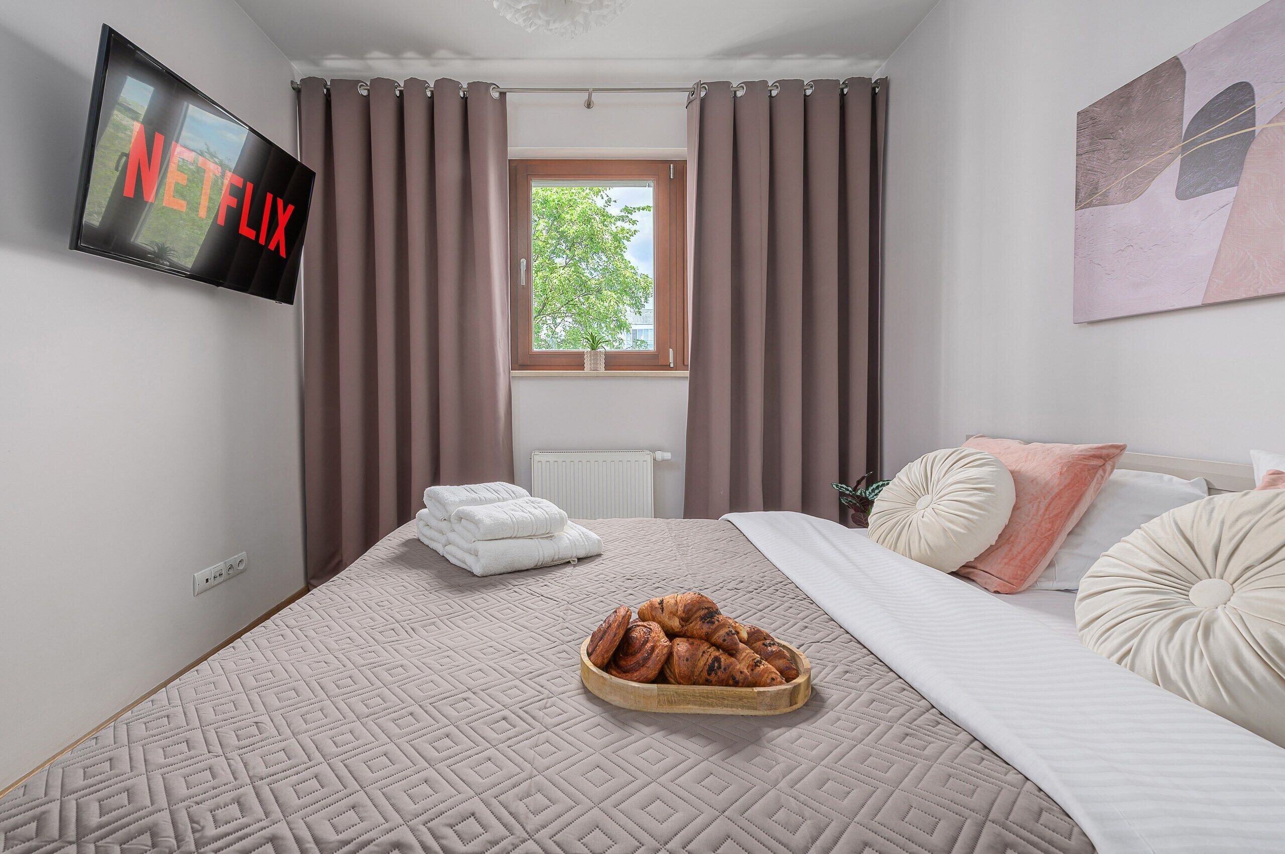 A hotel room with a bed, a TV mounted on the wall, a window with curtains, and a view of greenery outside.