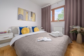 A neatly arranged bedroom with a large bed, a bedside table with books, a window with curtains, and a radiator. - Lumina 3-room Apart with Parking  (Warszawa)