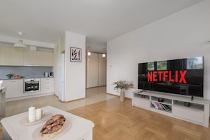 Ein modernes Wohnzimmer mit gro ßem Flachbildfernseher, der das Netflix-Logo zeigt, einer Küchenzeile mit weißen Schränken und einem Esstisch mit Brot.
