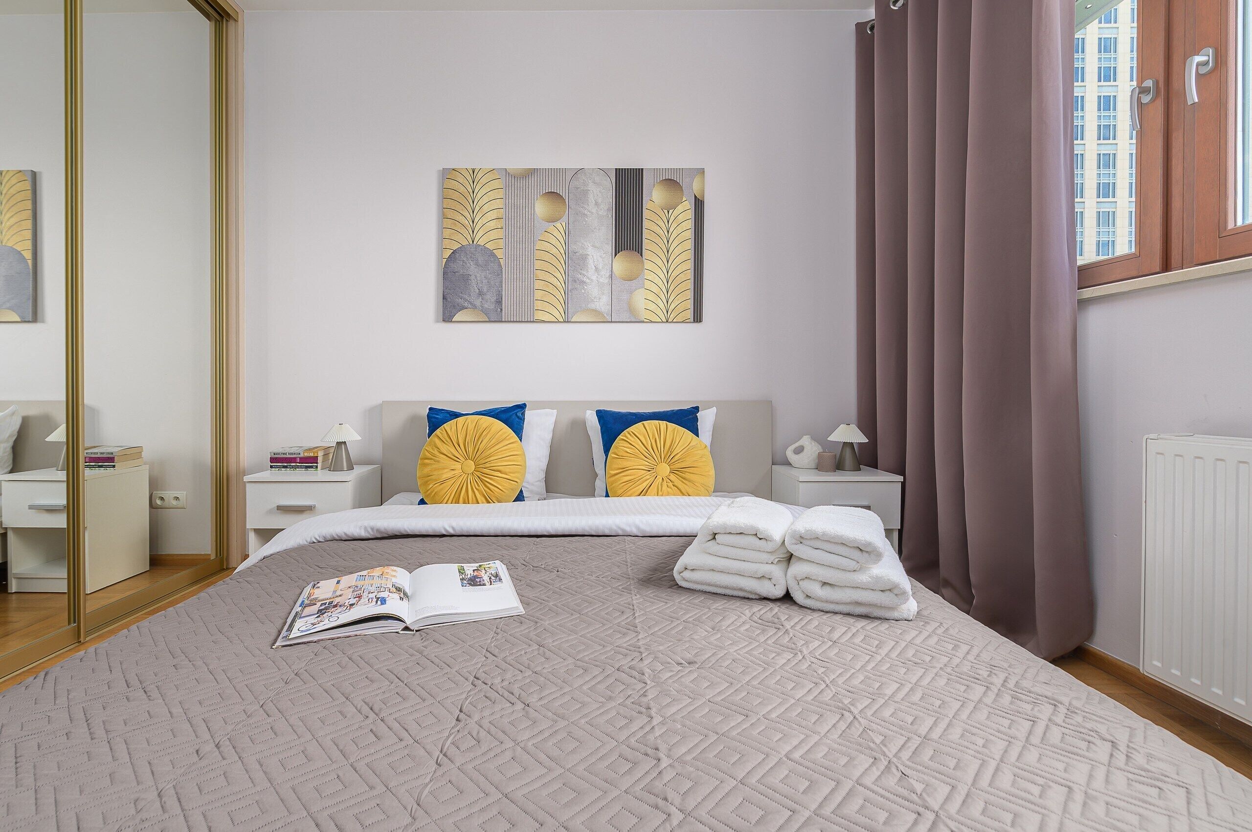 A hotel room with a bed, a bedside table, a lamp, a window with curtains, and a wall art piece.