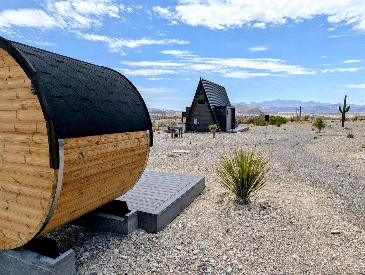A desert landscape with a large wooden barrel structure, a small building with a sloped roof, and a clear sky.