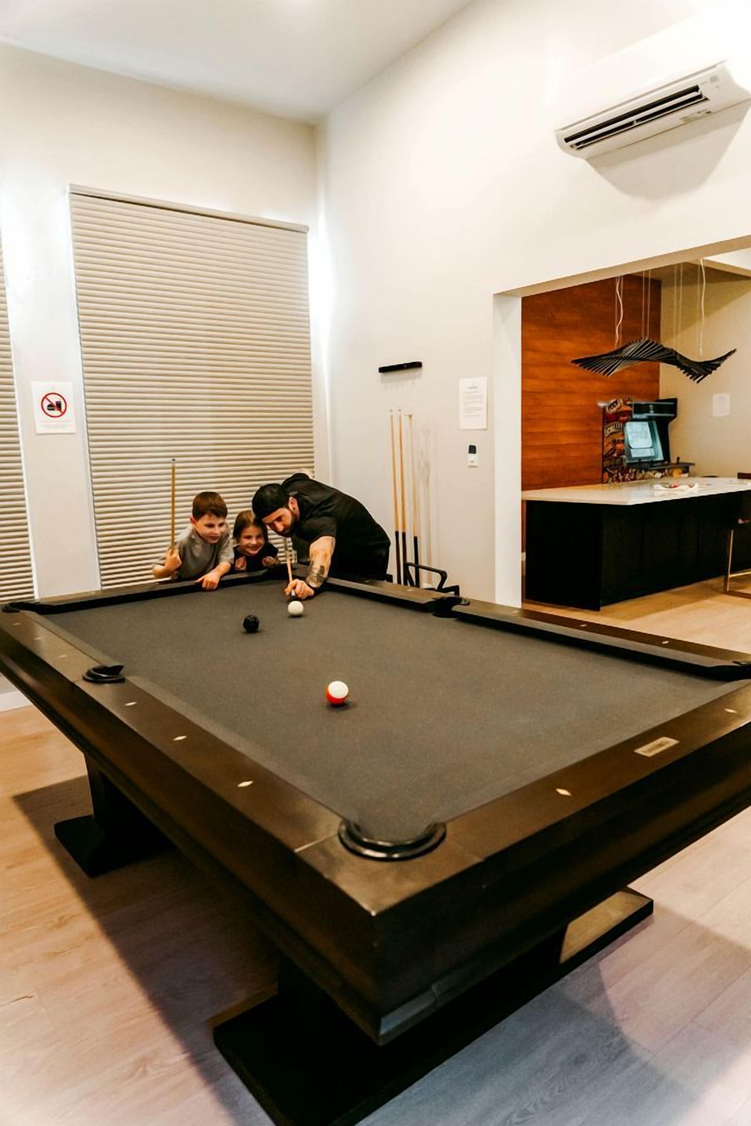 A pool table in a room with a closed door, a coffee machine, and a hanging light fixture.