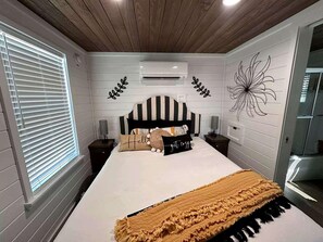 A bedroom with a large bed, a window with blinds, a wall-mounted air conditioner, and a decorative wall piece. - Charming Tiny Home Hideaway with Resort Pool Near Fredericksburg, TX (Fredericksburg)