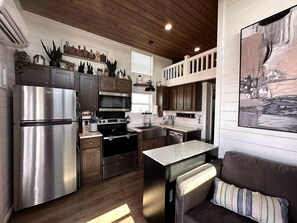 A modern kitchen with stainless steel appliances, wooden cabinets, and a dining area with a couch. - Charming Tiny Home Hideaway with Resort Pool Near Fredericksburg, TX (Fredericksburg)